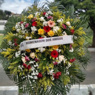 coroa de flores ricamente adornada. Ela apresenta uma mistura diversificada de flores, incluindo rosas amarelas, gerberas cor-de-rosa, flores brancas que se assemelham a crisântemos, além de flores vermelhas e roxas