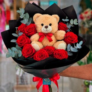 Arranjo romântico com ursinho de pelúcia de 23cm centralizado, cercado por 10 rosas vermelhas e ramos de eucalipto em embalagem de papel fosco preto com laço de cetim - Ikebana Flores BH.