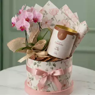 Box de presente cilíndrica com estampa floral contendo orquídea Phalaenopsis rosa, lata de biscoitos Delicê e laços de fita de cetim rosa seco.