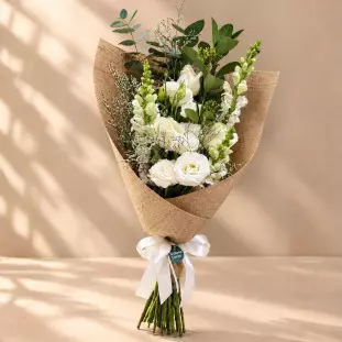 Buquê vertical com flores brancas variadas e folhagens verdes, envolvido em manta telada marrom e finalizado com laço branco
