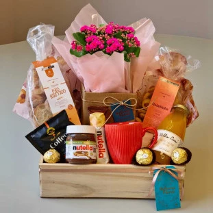 cesta de presente com uma planta de Kalandiva rosa envolta em embalagem plástica, decorada com um laço rosa. Ao lado, há outra parte da cesta envolta em tule rosa, contendo produtos diversos e outro laço rosa com uma etiqueta, simbolizando o carinho e del