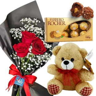 buquê com 3 rosas vermelhas rodeado de gipsonfila embalado por um papel preto e laço vermelho. Ao lado uma caixa de chocolates ferrero rocher e um ursinho de pelúcia.