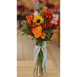 Aura Alegre em vaso de vidro, com girassol, rosas cor-de-rosa, alstroemérias vermelhas e mini rosas laranja, em estilo vibrante e alegre.