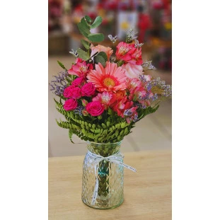 Arranjo Aura Rosada com flores em tons de rosa e pêssego, incluindo gérbera, rosas e alstroemérias, em vaso de vidro transparente decorado com laço.