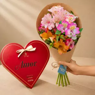 Kit de presente com caixa em formato de coração vermelho escrito Amor com biscoito Red Velvet e um buquê pequeno de astromélias coloridas em papel kraft sendo segurado por uma mão - Ikebana Flores BH.