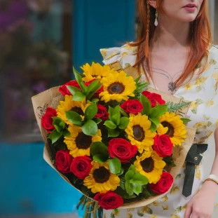 Mulher segurando buquê extra grande com 12 rosas vermelhas, 8 girassóis e folhagens em embalagem de jornal.