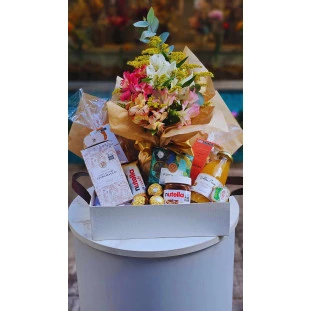 Cesta de café da manhã com flores de astromélias e produtos gourmet como Nutella, Ferrero Rocher e suco natural.