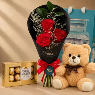 buquê com 3 rosas vermelhas embalado por um papel preto e laço vermelho. Ao lado uma caixa de chocolates ferrero rocher e um ursinho de pelúcia.