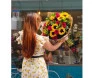 Mulher de costas segurando buquê extra grande com 12 rosas vermelhas, 8 girassóis e folhagens em embalagem de jornal.