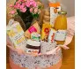 cesta de presente com uma planta de Kalandiva rosa envolta em embalagem plástica, decorada com um laço rosa. Ao lado, há outra parte da cesta envolta em tule rosa, contendo produtos diversos e outro laço rosa com uma etiqueta, simbolizando o carinho e del