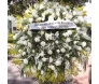 A imagem mostra uma bela coroa de flores predominantemente branca. As flores brancas, possivelmente rosas, lírios e pequenas margaridas, são dispostas de forma densa e luxuriante, intercaladas com verdes folhagens que proporcionam contraste e profundidade