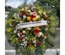coroa de flores ricamente adornada. Ela apresenta uma mistura diversificada de flores, incluindo rosas amarelas, gerberas cor-de-rosa, flores brancas que se assemelham a crisântemos, além de flores vermelhas e roxas